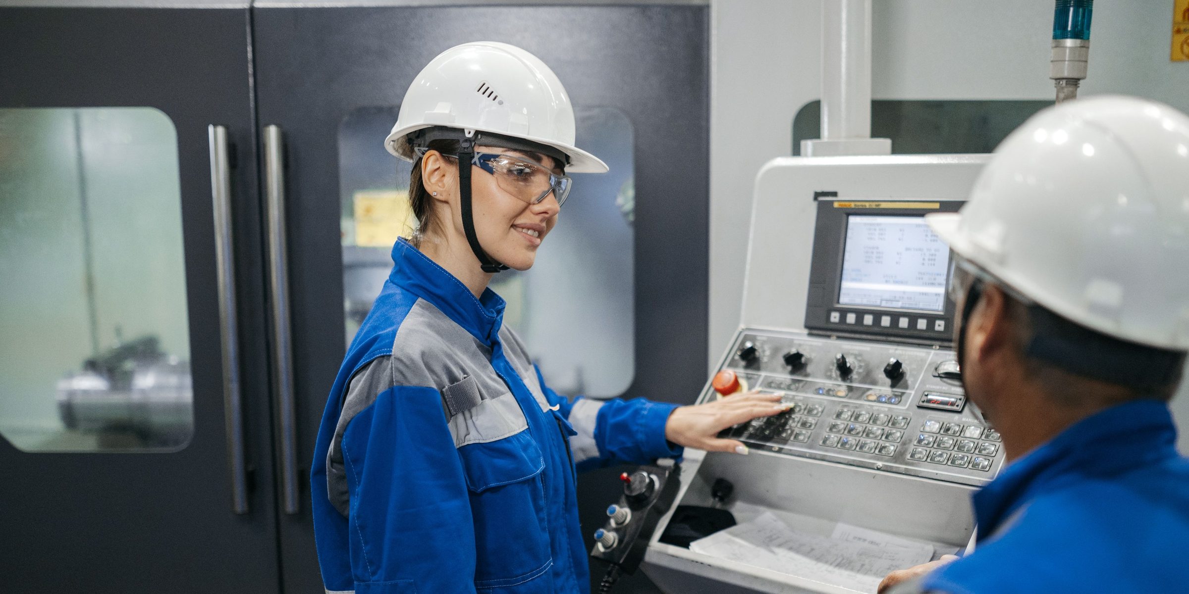 An operator in a hard hat and safety glasses runs a CNC machine control panel while a colleague looks on